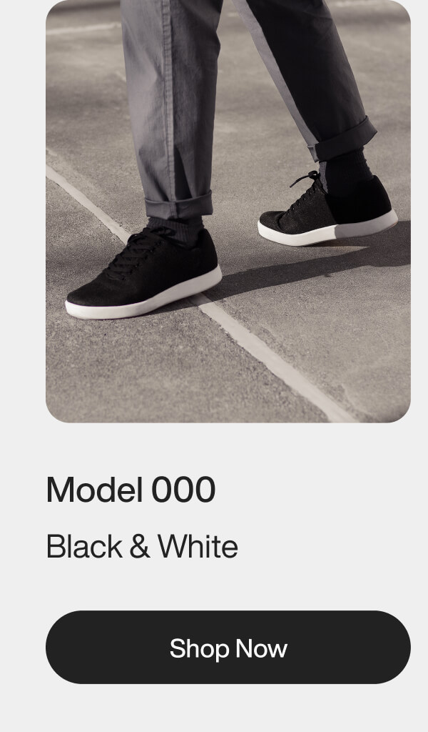 A person wearing gray pants and black sneakers with white soles stands on a pavement. Text below reads: "Model 000, Black & White, Shop Now."