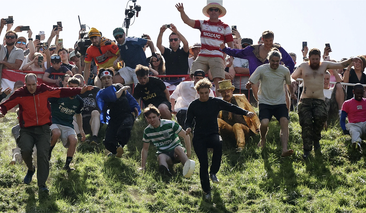 A crowd of people chase a cheese wheel down a hill.