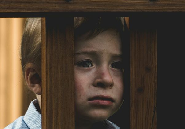 A young child with blonde hair peers through the vertical slats of a wooden railing, their expression somber and tearful.