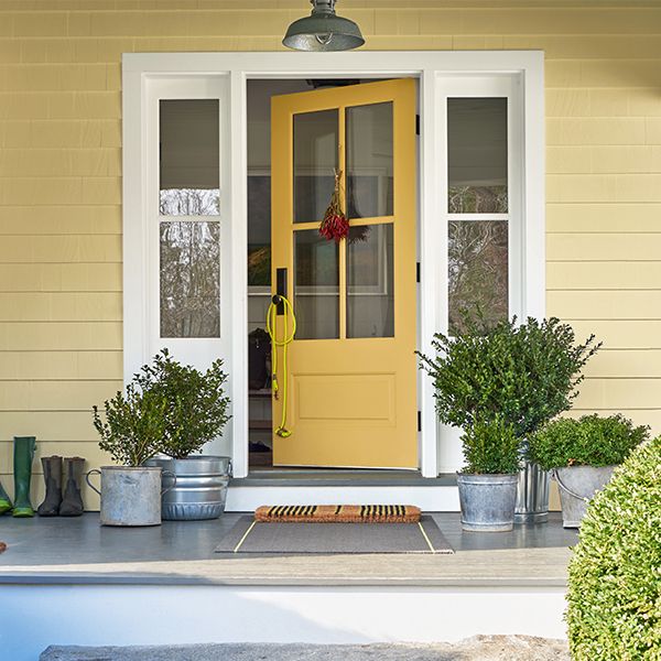 The front porch of a yellow-painted house with a yellow door, white trim, plants in metal pails, and two round shrubs out front.