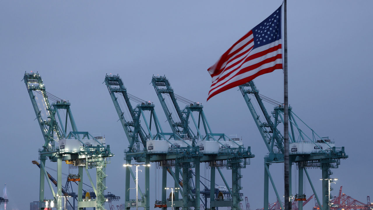A view of shipping cranes at the Port of Los Angeles in San Pedro, California - with a US flag waving in the foreground.