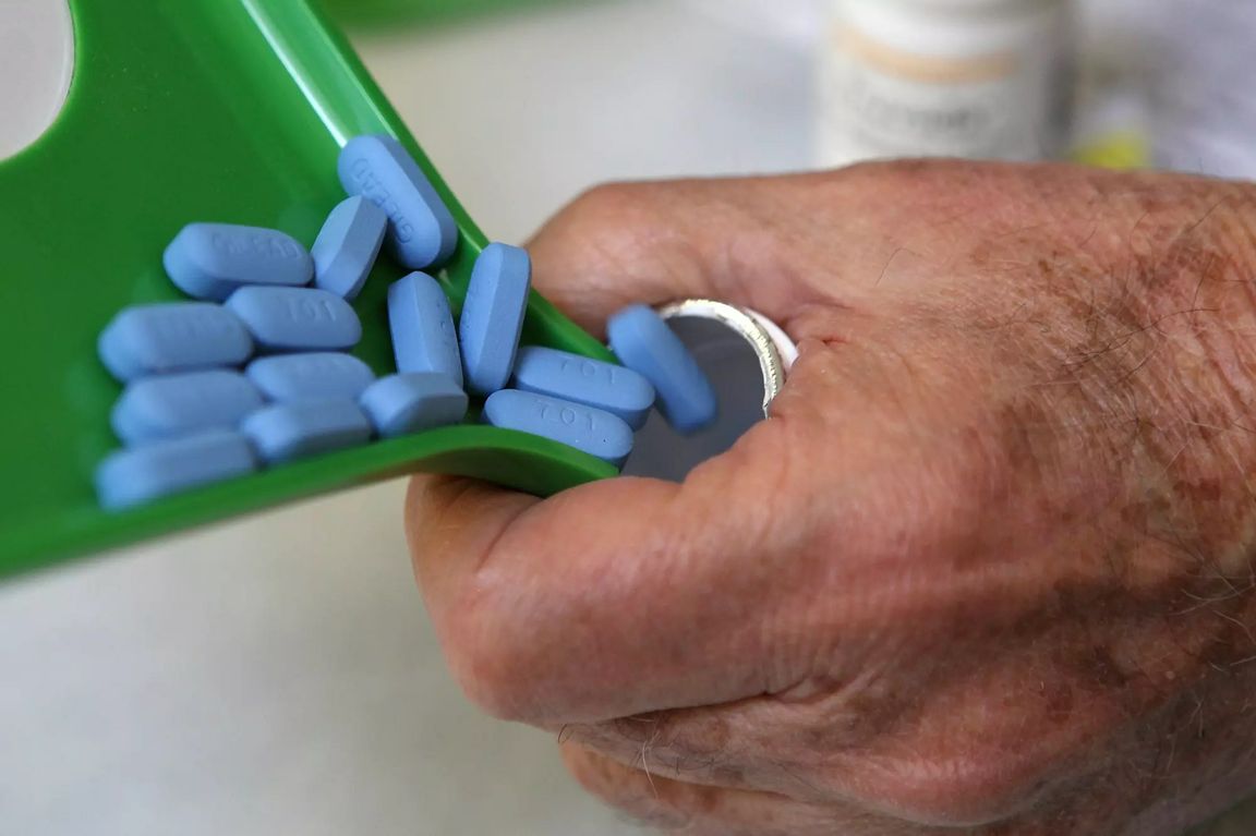 A close-up shows blue prescription tablets being poured from a green pill tray into a person’s hand.