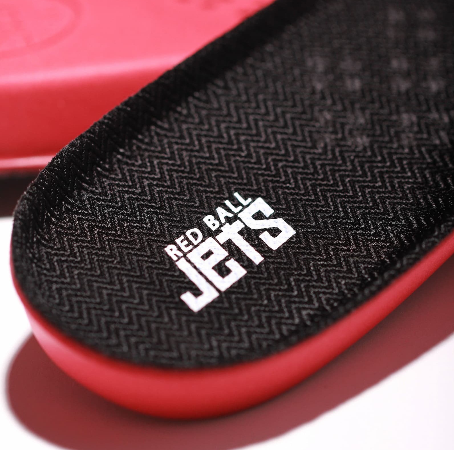 Close-up of a Red Ball Jets insole showing the textured black fabric top surface with a chevron weave pattern and embossed white Red Ball Jets logo. The red PU base of the insole is visible along the edge.