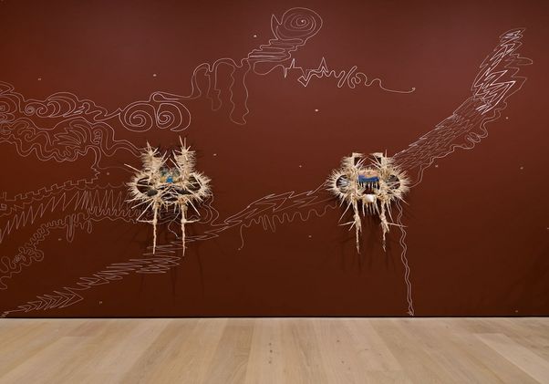 Wall installation with two straw-and-fiber sculptural forms mounted on a reddish-brown wall, connected by white, hand-drawn abstract lines resembling swirling landscapes, waves and lightning across the surface.