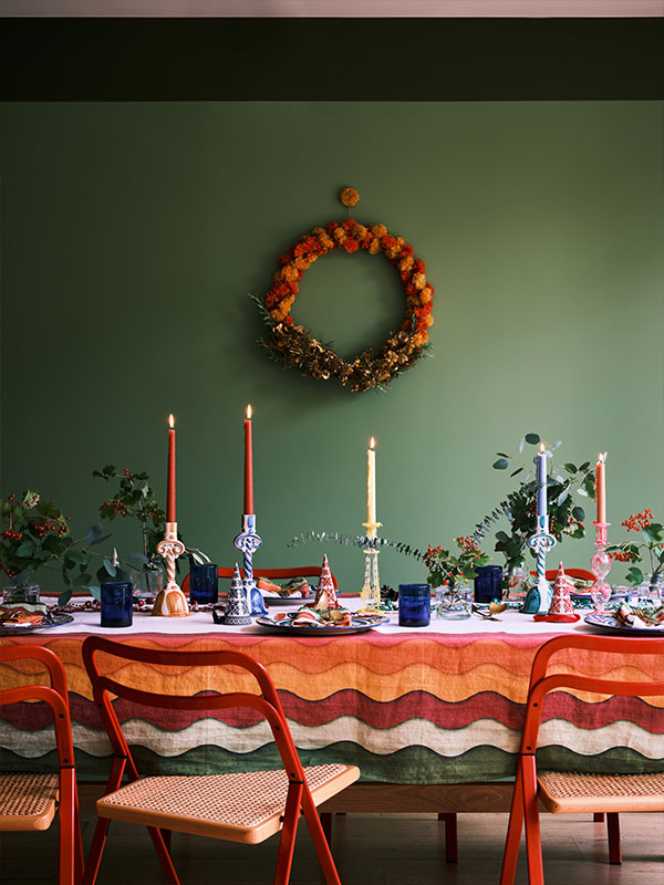 A festive dining table with lit candles, colourful wavy tablecloth, and a floral wreath on a green wall.