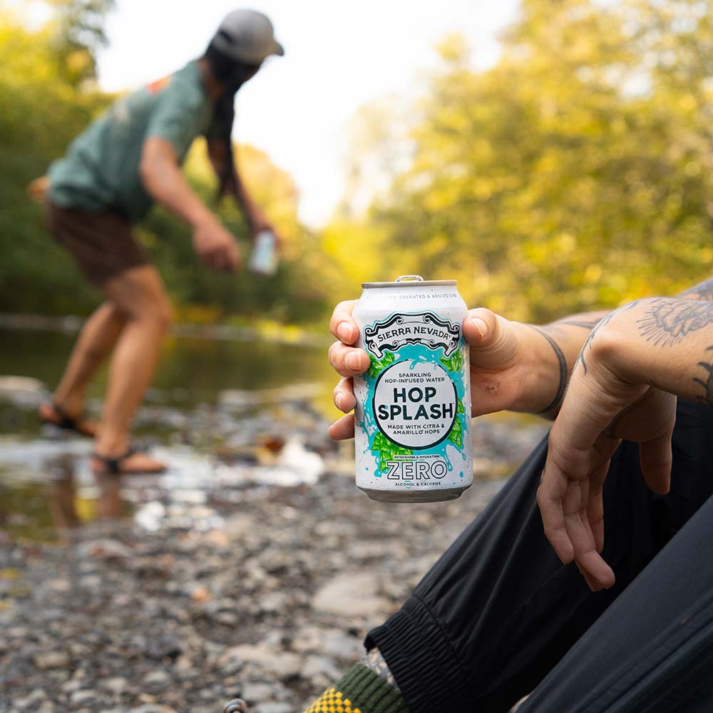 A hand holding a Hop Splash can in the foreground, and a person skipping rocks on a creek in the background