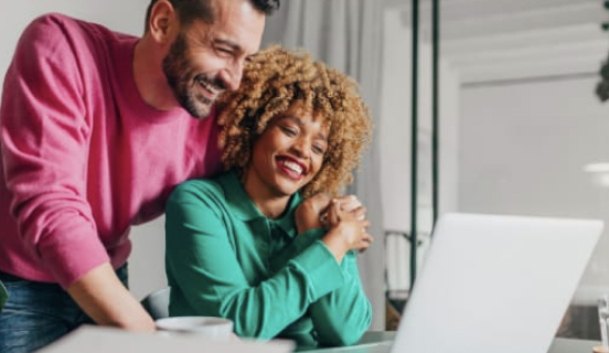 A smiling man and woman stare at an open laptop.