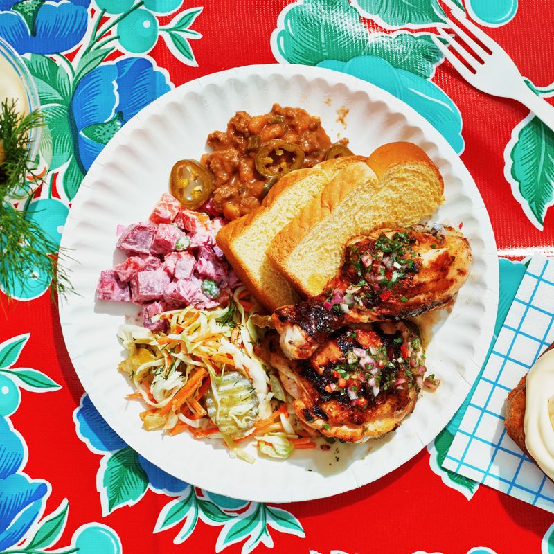 Pink potato salad, pickle coleslaw, burger beans, better-than-beer-can chicken, banana pudding cake, and slices of bread on a plate with a glass of pickle lemonade.