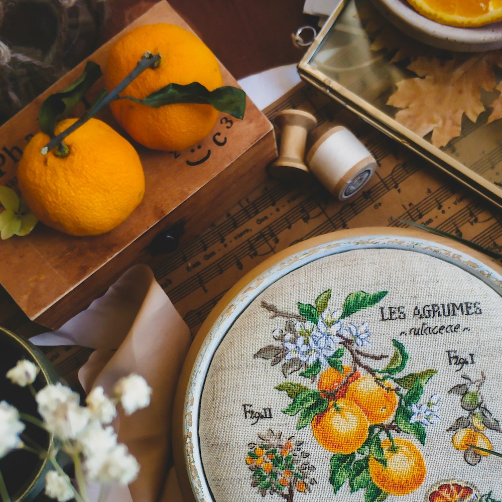 An embroidery of clementines growing on a branch. Surrounding the embroidery hoop are real-life clementines, sheet music, and spools of thread.