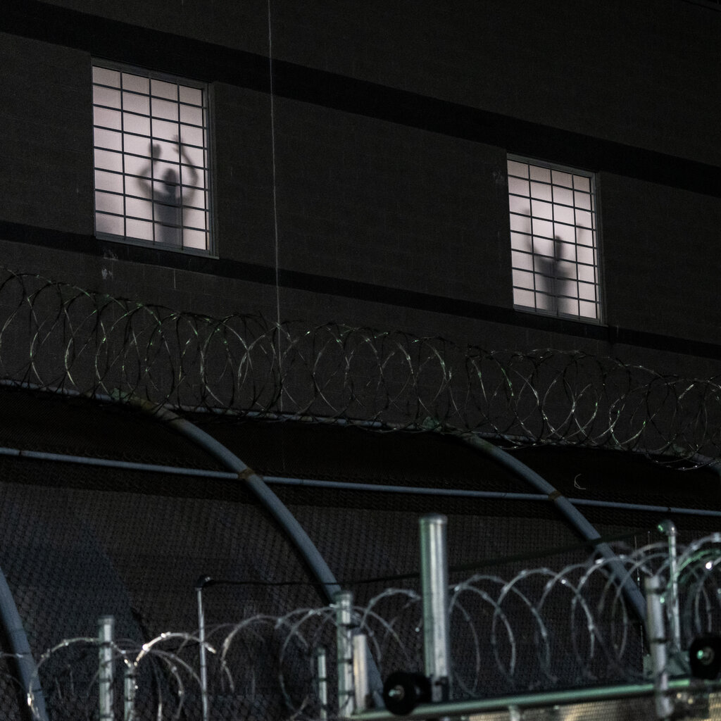 A dark building’s three lit windows show silhouettes of people behind grates. Razor wire is in the foreground.