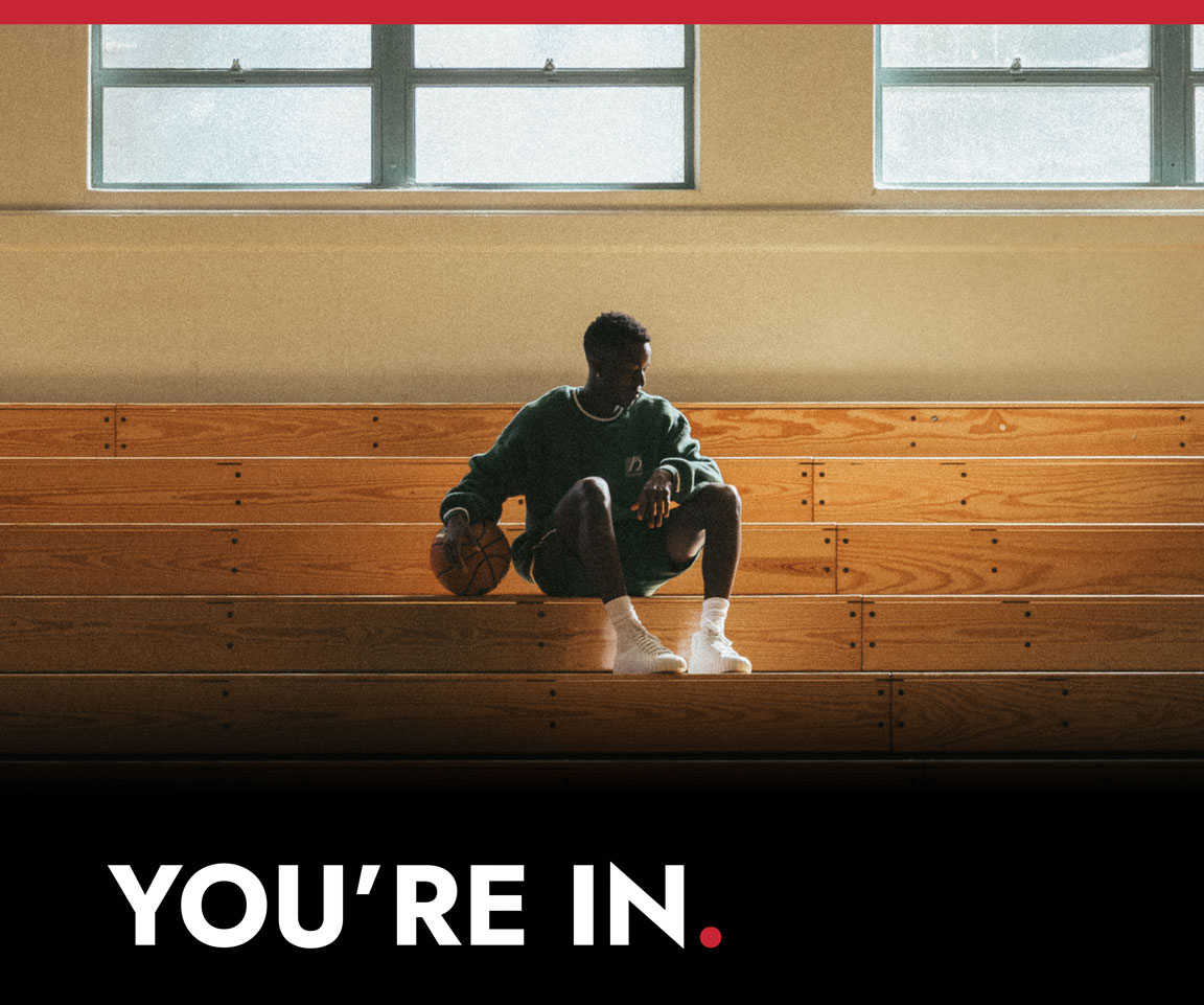 A person sits alone on wooden bleachers in a sunlit gym, holding a basketball and wearing white Red Ball Jets. Bold text at the bottom reads, “YOU’RE IN.”