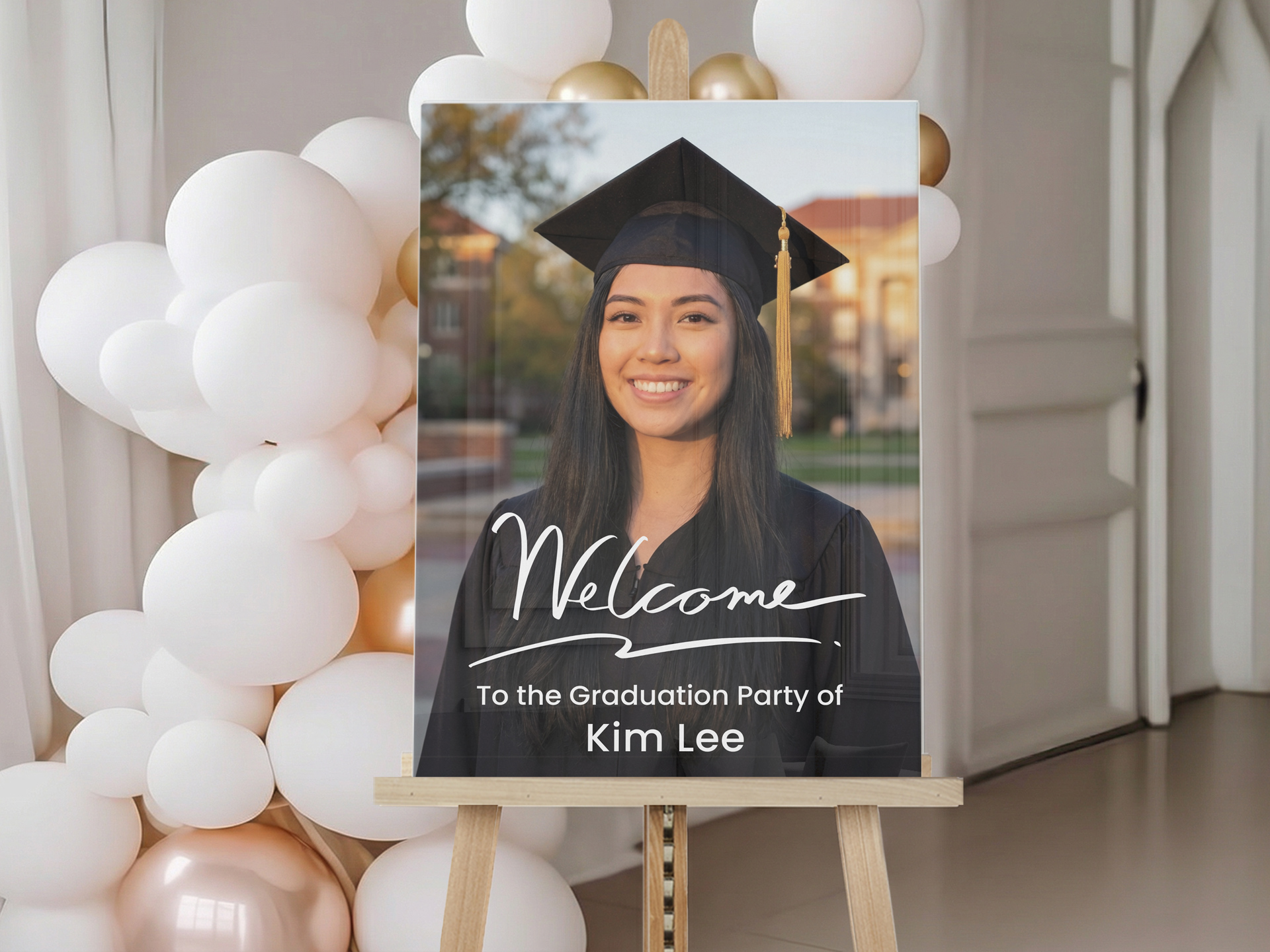Graduation welcome sign on an easel featuring a graduate portrait and “Welcome to the Graduation Party of Kim Lee” text, displayed beside white and gold balloons.