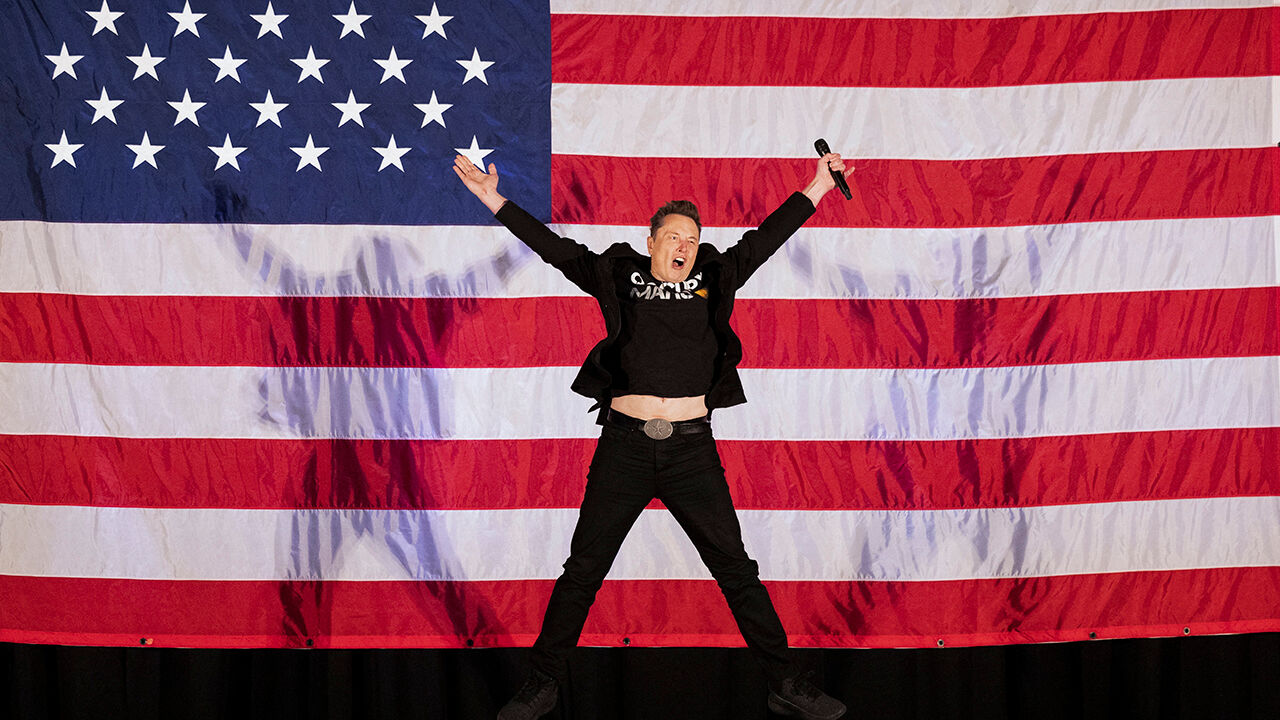 Elon Musk jumps up and down on a stage in front of the American flag