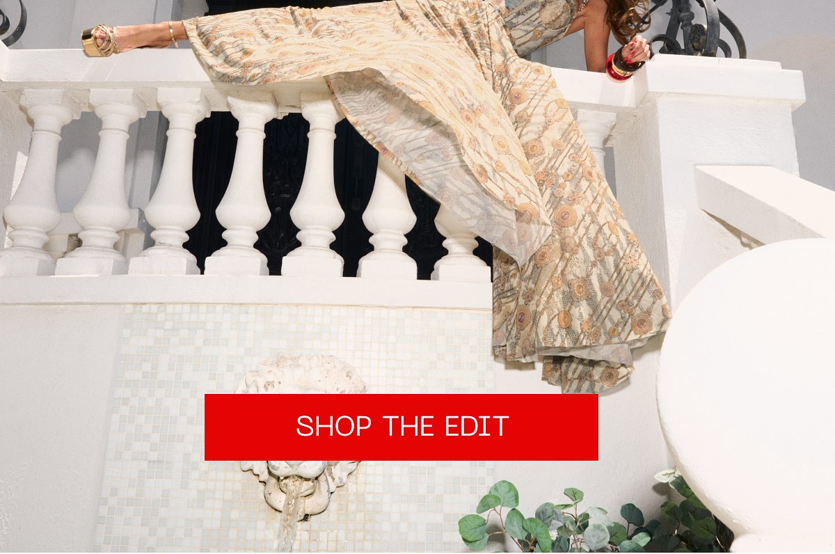 A model in a flowing dress poses on a balcony with a fountain.