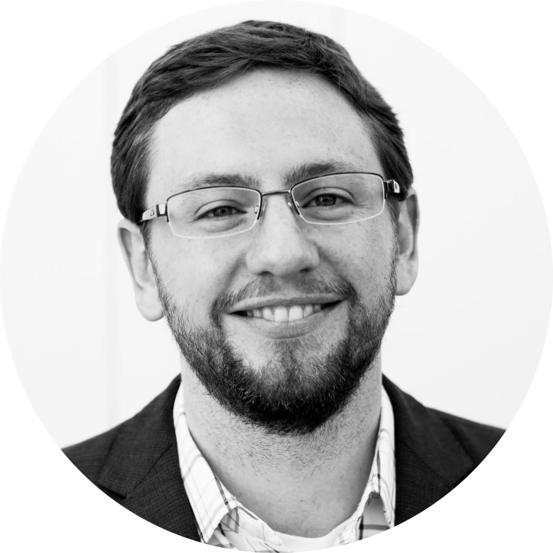 Dylan, a white man with black glasses, neat swept-back hair and a short beard, smiles, sporting a dark suit.