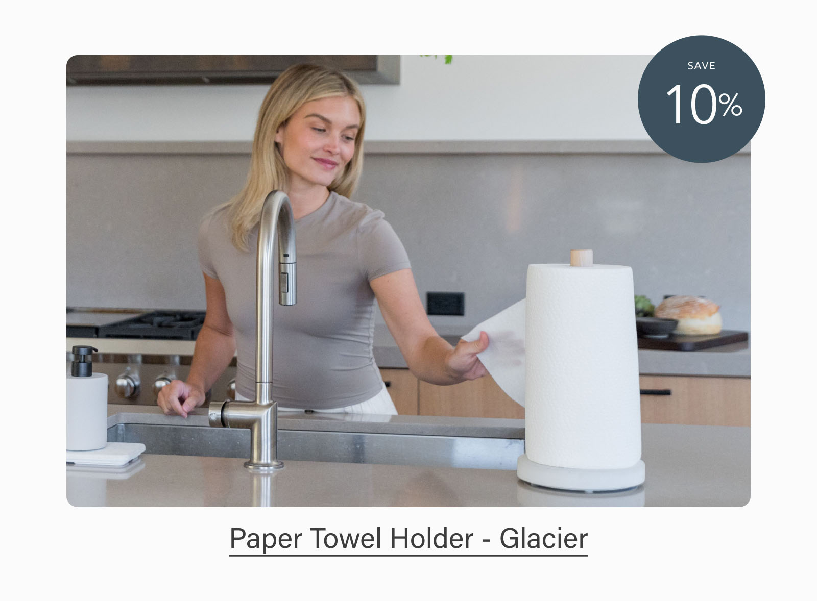 Woman reaching for a paper towel from a white Dorai Paper Towel Holder with a stone base on a kitchen countertop. Badge indicates 10% savings. Text reads: "Paper Towel Holder - Glacier."