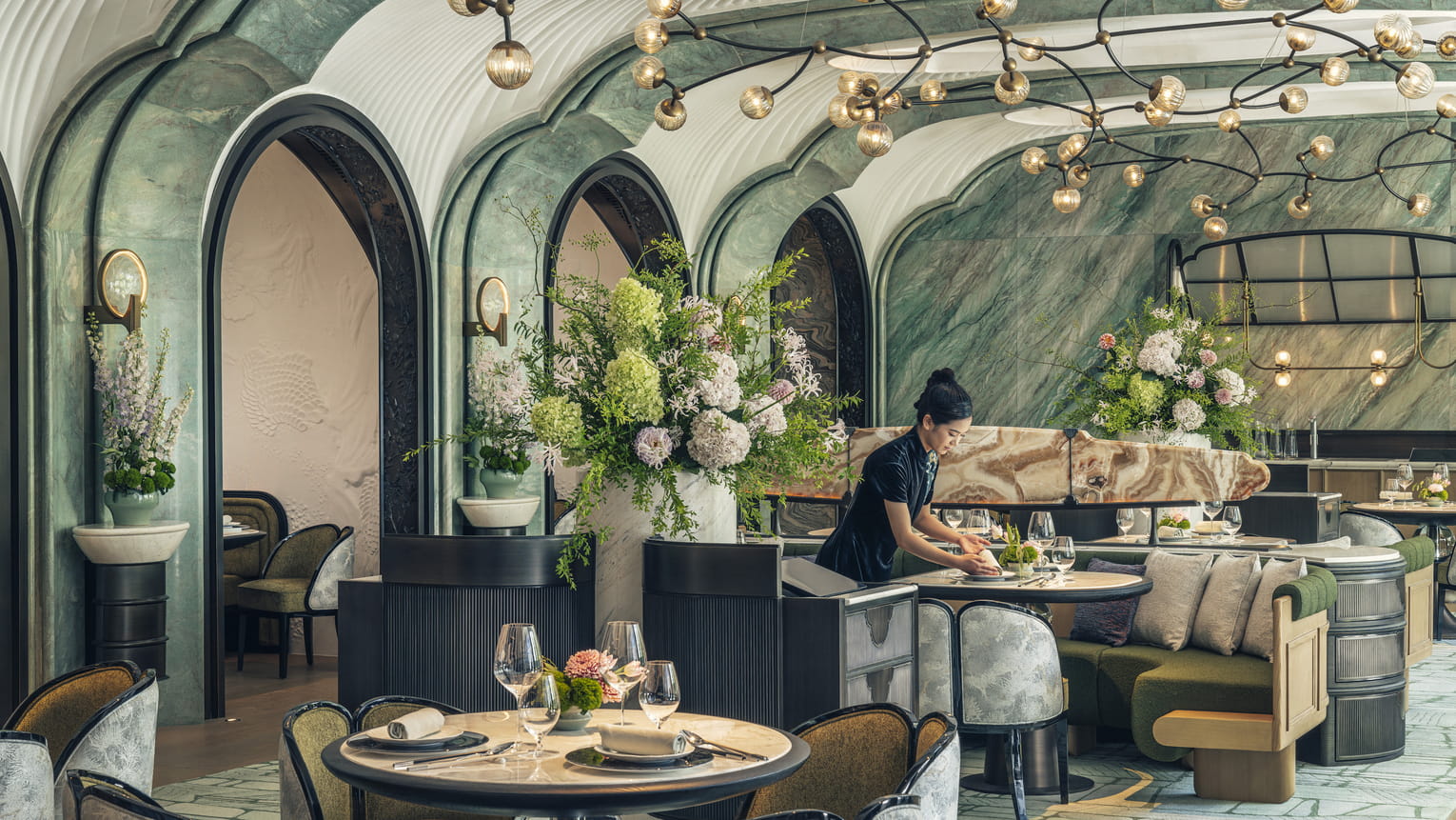 A couple at a restaurant watching as a sous chef pours the finishing sauce on an artfully prepared dish.