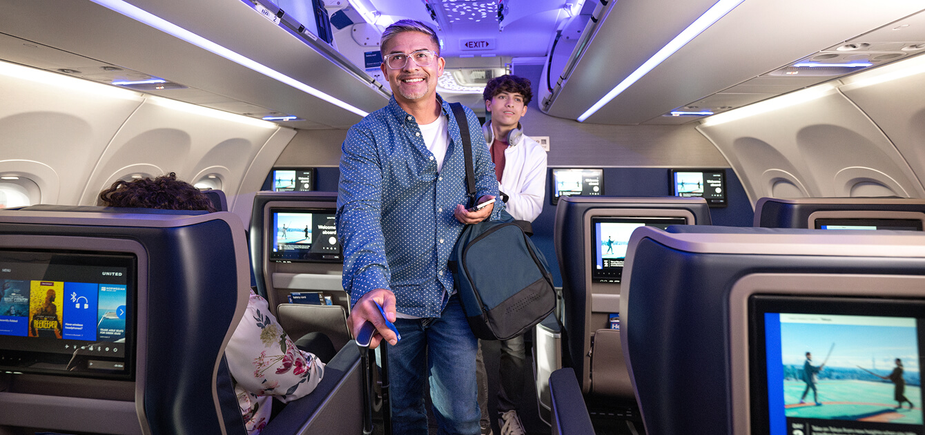 Two smiling men in the aisle of an aircraft with their carry-on luggage.