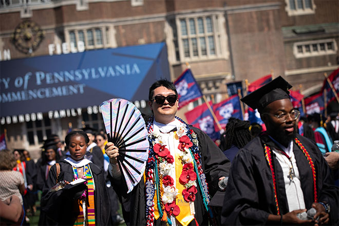 A student in a graduation robe holding a fan and wearing flower necklaces.