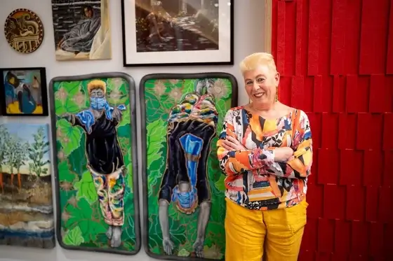 A person stands in front of their artwork as they smile at the camera with their arms crossed over their chest.