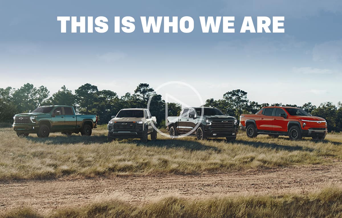 This is who we are. A lineup of four Chevy trucks parked off-road in a field with trees in the background.
