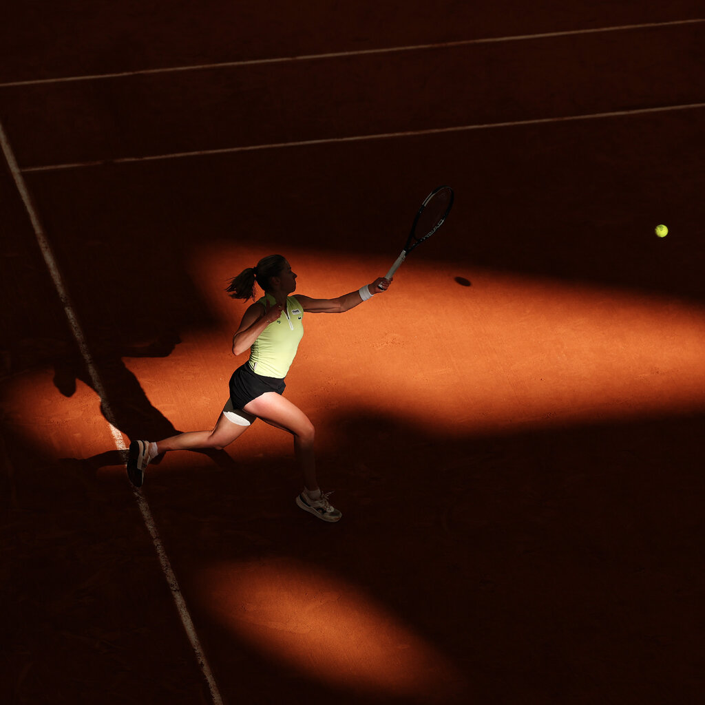 A person in a light green top hits a yellow tennis ball on a clay court. Half the court is covered in a deep, dark shadow.