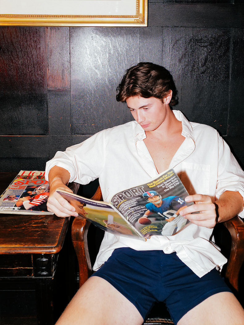 Man sitting reading a football magazine and he is wearing white dress shirt and blue slim fit boxers