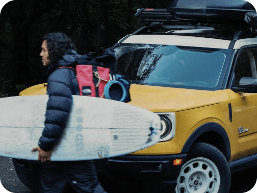 A man wearing a backpack and carrying a surfboard in front of a parked Bronco&reg; Sport SUV
