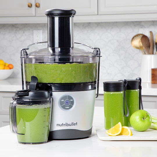 A nutribullet Juicer Pro™ with green juice sits on a kitchen counter. Nearby are a pitcher, two bottles, and a plate with an apple and lemon wedges.