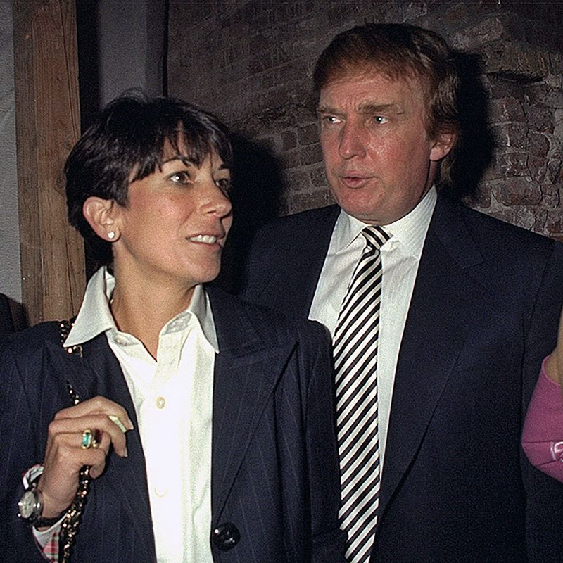 Two figures stand close together. On the left a woman with short hair in a blazer looks to another figure. On the right another wears a suit and tie and is speaking, looking towards the woman. 