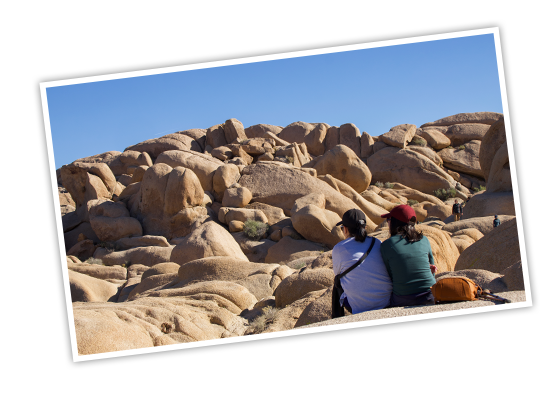 Two hikers sit on a boulder and look out at the many smooth, boulders in front of them that form a small mountain at Joshua Tree National Park