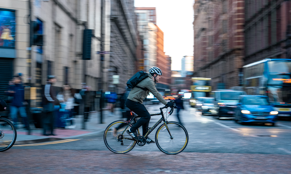 A commuter crosses a busy city-centre junction on his road bike with traffic queueing in the background.