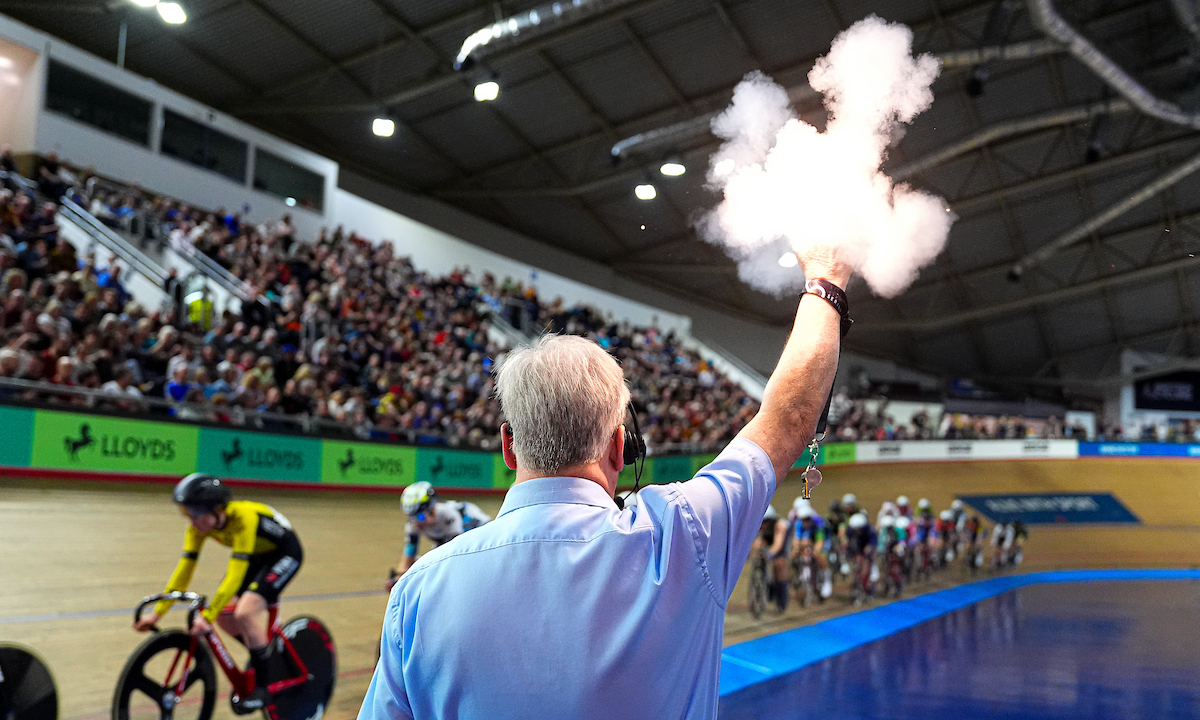 A commissaire wearing a blue shirt fires the starting pistol for a track event at the National Cycling Centre, with riders and the crowd in the background.