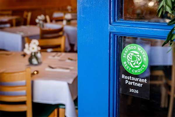 A green Seafood Watch Best Choice Restaurant Partner 2026 decal on the window pane of a blue door opening to restaurant seating