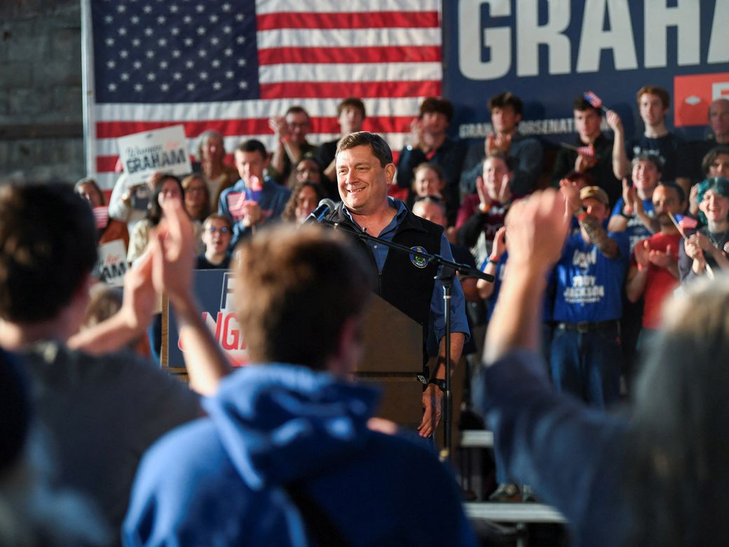 Figures rally around another who stands at a podium on stage.