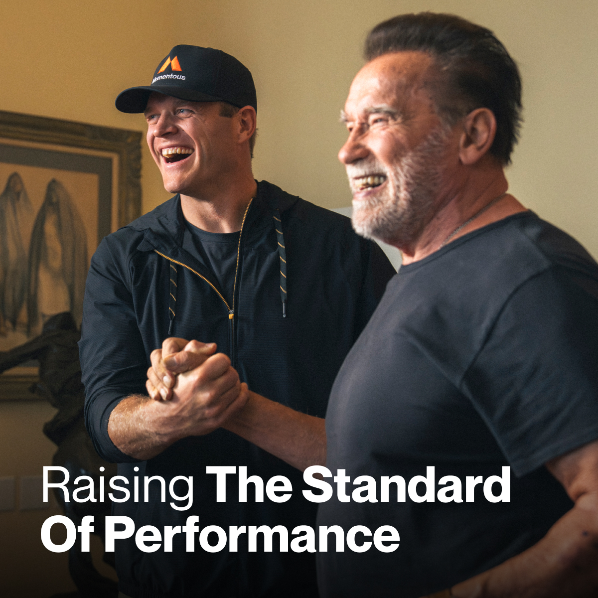 Arnold Schwarzenegger and Jeff Byers standing indoors, smiling and clasping hands. One is Arnold Schwarzenegger wearing a black T-shirt; the other wears a black jacket and Momentous hat. White text reads, “Raising The Standard Of Performance.”