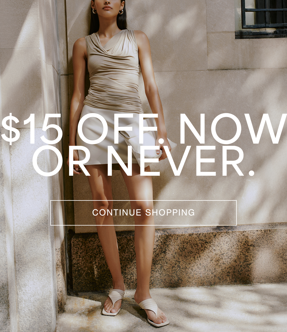 Sarah stands against a sunlit stone wall wearing the Sutton Top in sand, styled with a white mini skirt and sandals. Overlay text reads: “$15 OFF. NOW OR NEVER.” with a “CONTINUE SHOPPING” button beneath.