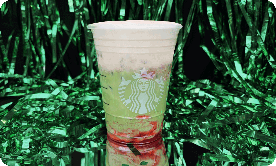 Iced green drink with red fruit on the bottom and light pink cold foam in a clear Starbucks to-go cup sits on a reflective surface in front of a green tinsel background.