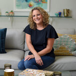 Jill Smokler sits on a couch, smiling for the camera. She has curly hair and is wearing jeans.