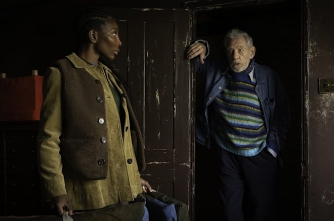 Michaela Coel stands in a dark wood room, looking at Ian McKellen, who holds a door open into a shadowed space.