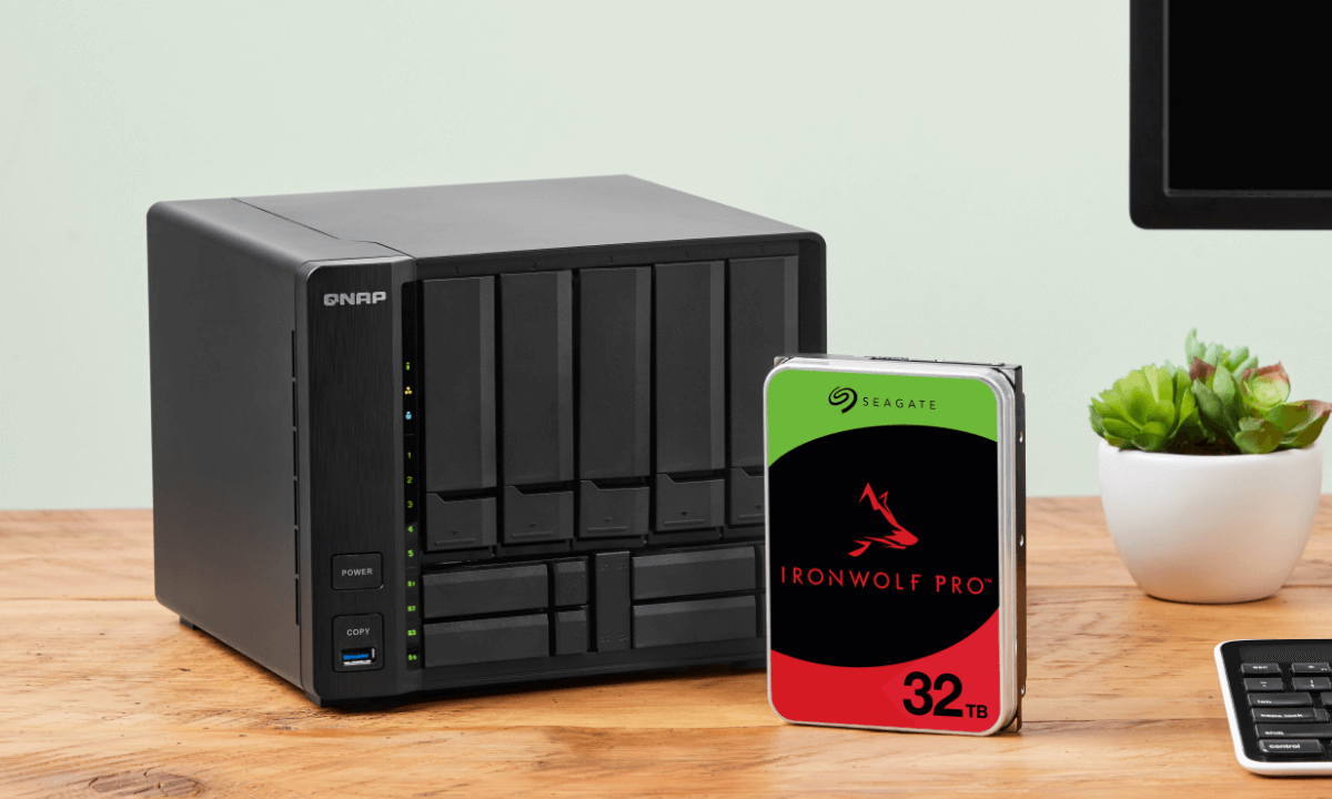 A black QNAP storage device sits on a wooden desk beside a Seagate IronWolf Pro 32TB hard drive, with a small potted plant nearby.
