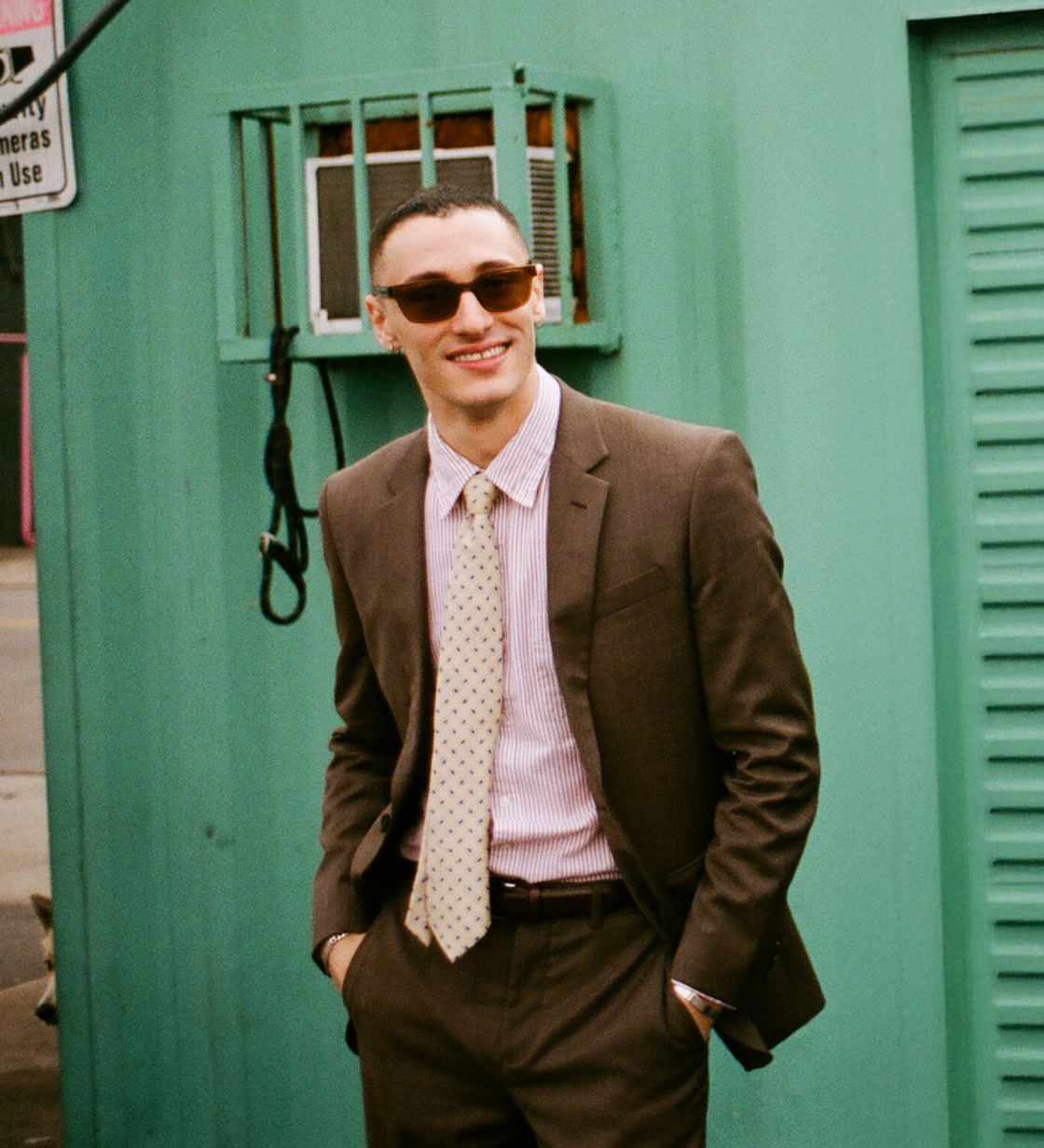 Man outdoors wearing a brown suit with striped shirt, patterned tie, sunglasses, and smiling with hands in pockets.