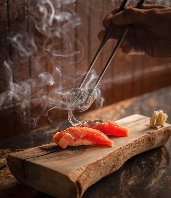 Chef sears sushi with smoking charcoal over a wooden board.