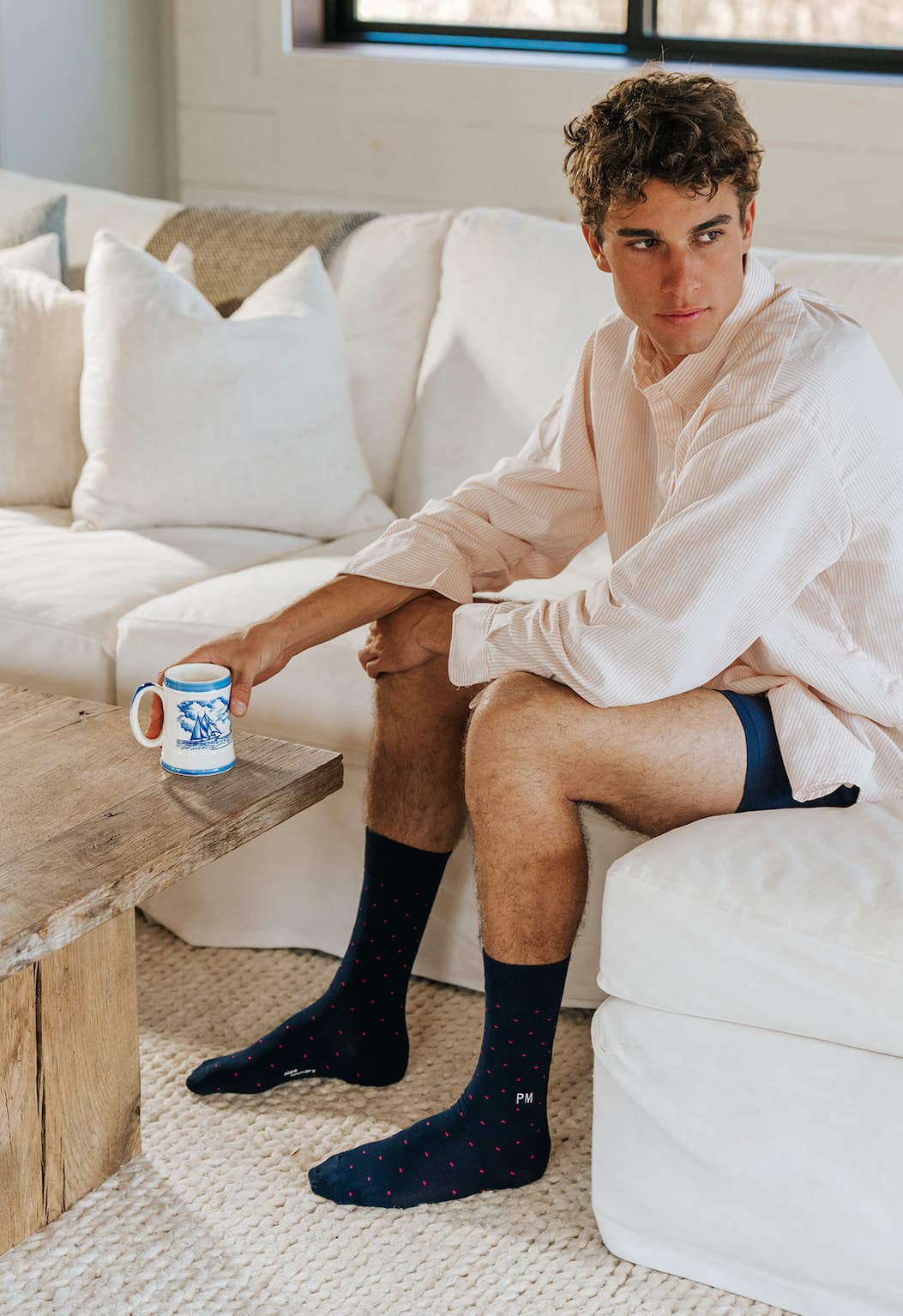 Man sitting on couch drinking coffee wearing white dress shirt and blue dress socks with letters PM monogrammed 