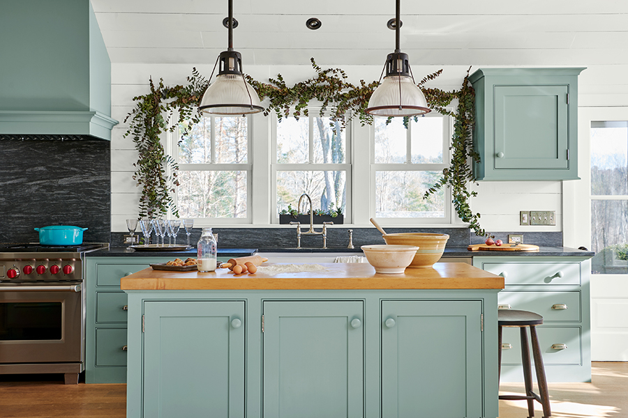 A kitchen with teal-painted cabinets, a larger butcher block island, and holiday swag draped over the windowsill.