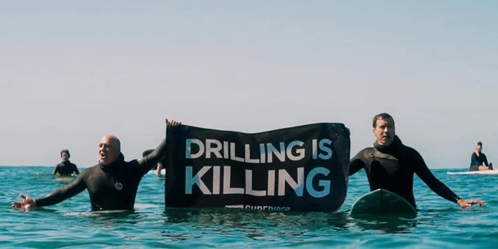 Two surfers out in the water hold a sign that reads, "Drilling is Killing."