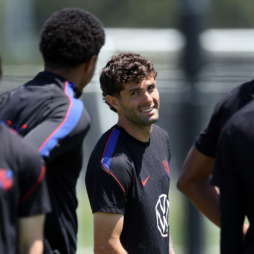 The soccer player Christian Pulisic, wearing a black jersey, smiles at the camera.
