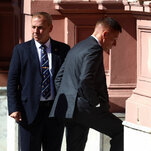 A person in a dark suit and blue tie stands next to another person in a dark suit stepping up white stairs. A reddish-brown building is in the background.
