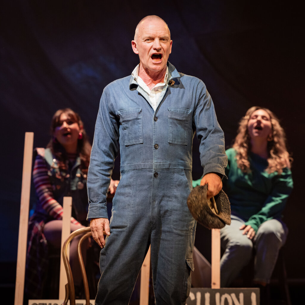 Sting is singing onstage, wearing a blue jumpsuit and holding a cap in his hand. Two performers are seated and singing behind him. 
