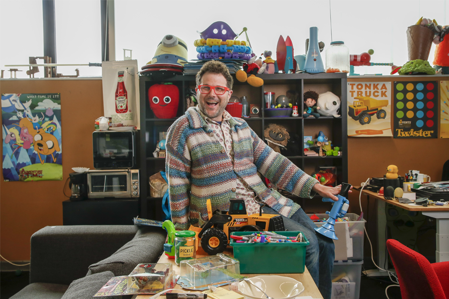 A man smiling sitting on a table in his office that's decoratived with colorful knickknacks.